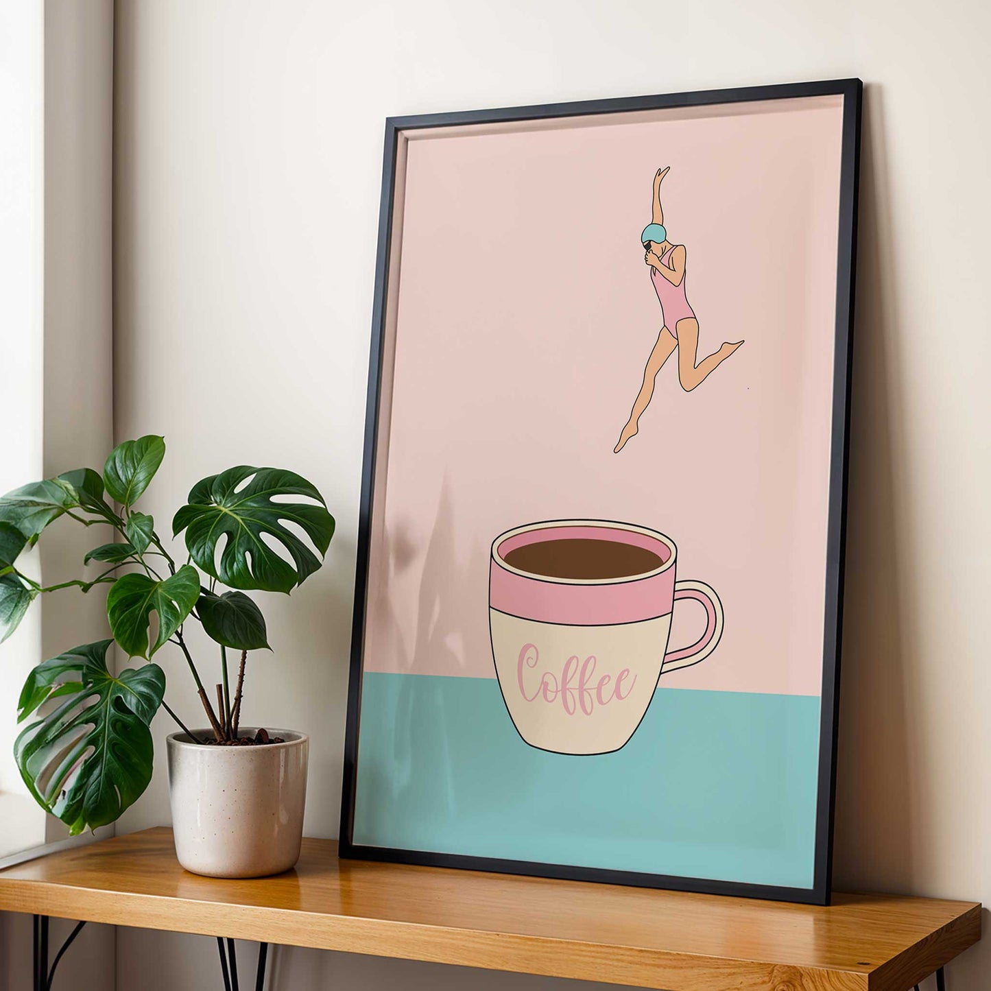 Framed artwork of a woman leaping over a coffee cup on a pink and blue background, placed on a wooden table with a plant.
