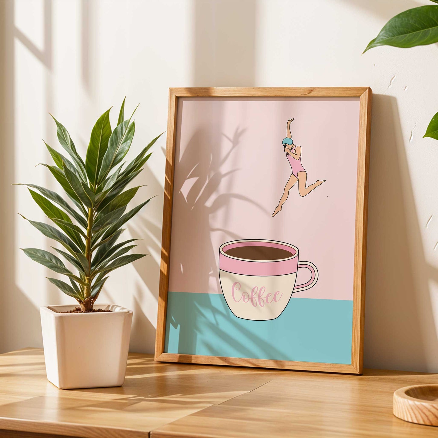 Framed artwork of a coffee cup with a person leaping over it, placed on a wooden surface with a plant in the background.